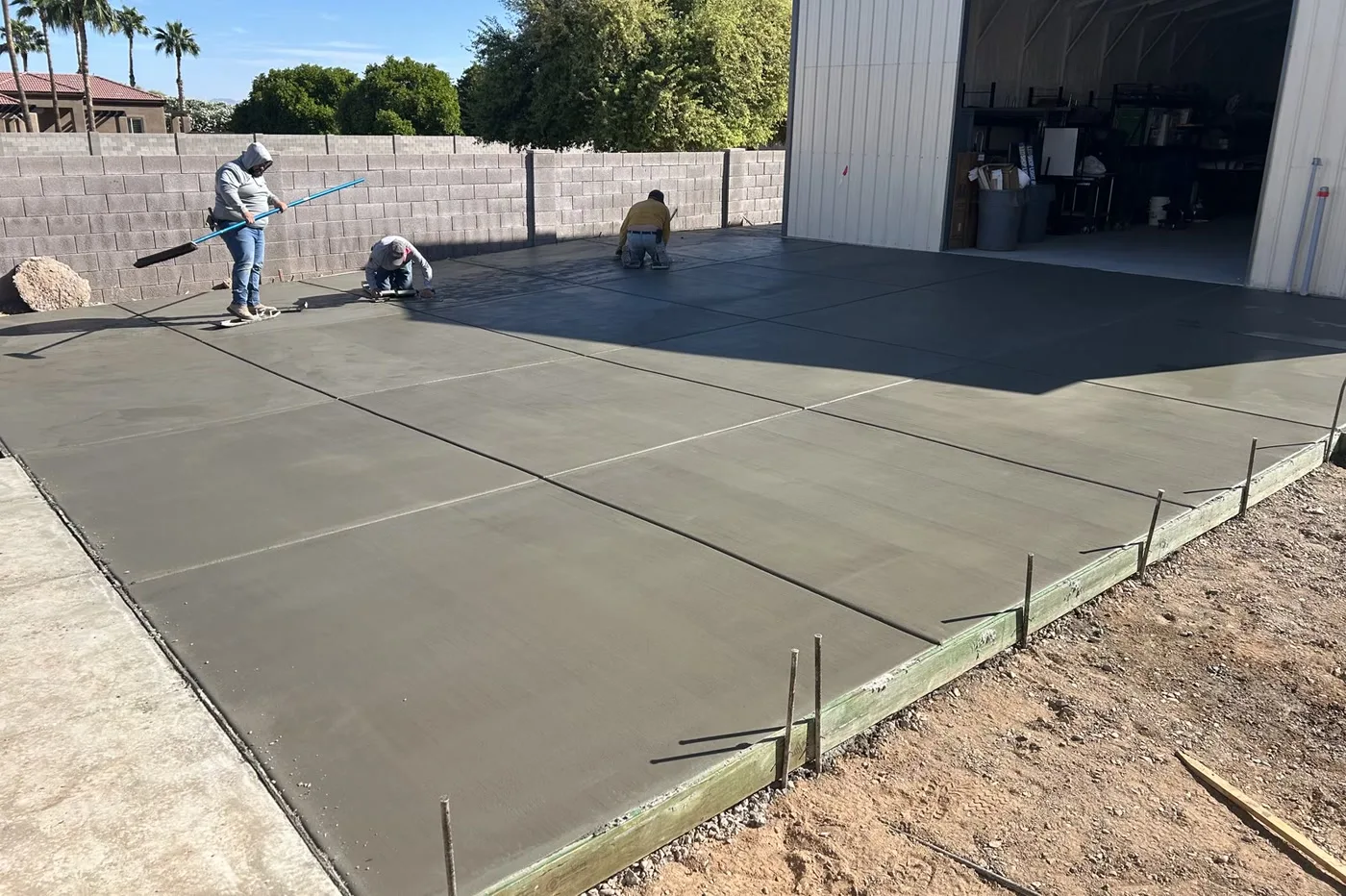 Crew finishing a freshly poured concrete pad at a steel building with the roll-up door open